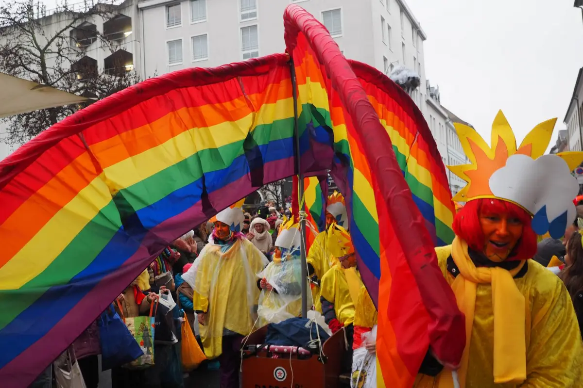 Bunter Karnevalsumzug mit großem Regenbogen-Banner, verkleideten Teilnehmern in gelben Kostümen und Sonnendekoren