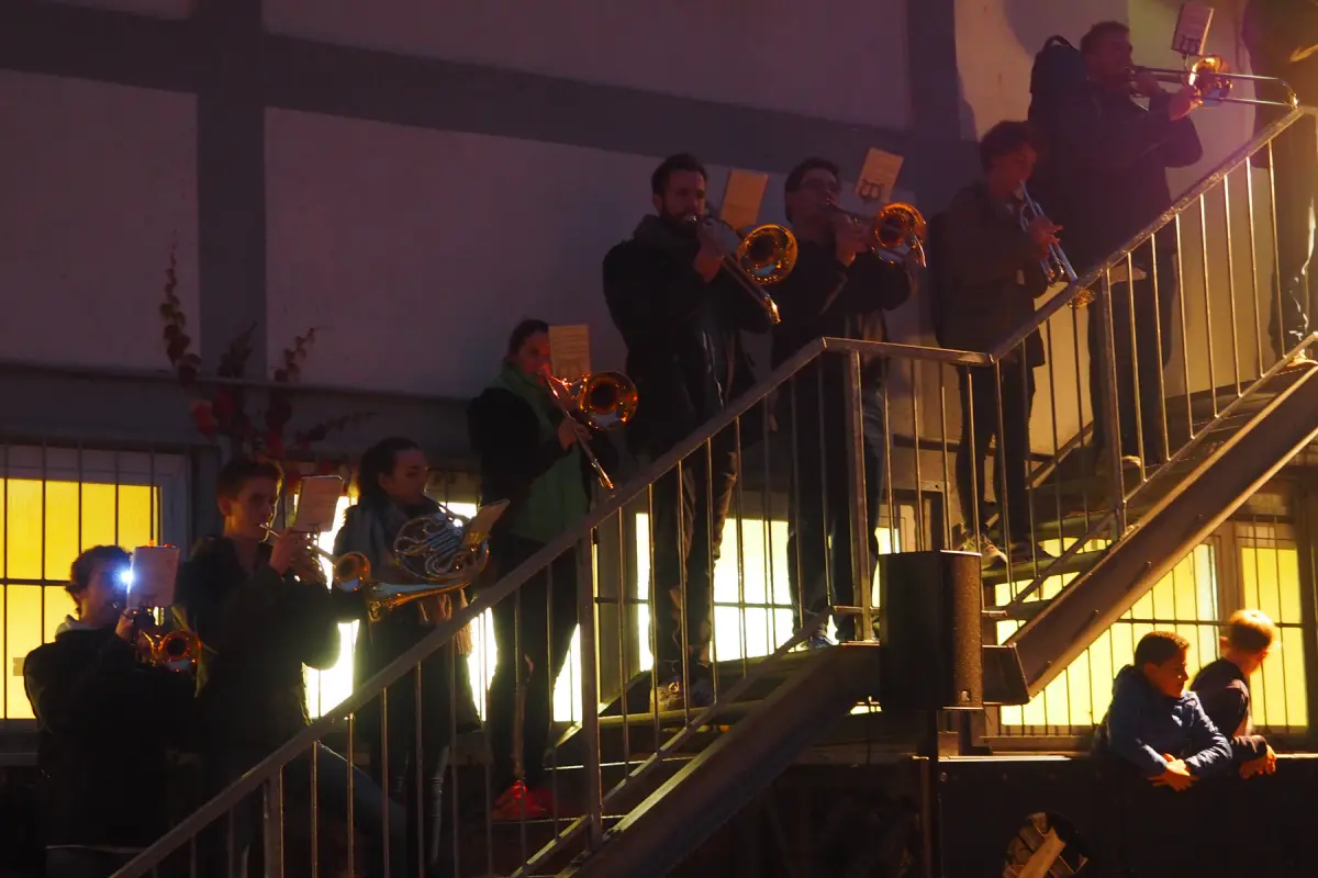 Bläserensemble spielt auf einer beleuchteten Außentreppe bei Dämmerung, zwei Zuschauer rechts sitzend