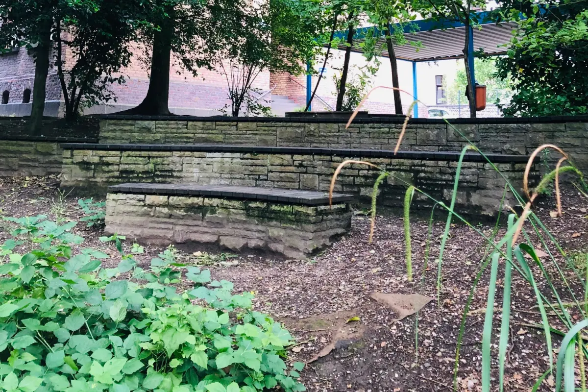 Terrassenförmige Natursteinstufen unter dichtem grünen Baumdach, Sträucher im Vordergrund und blaues Vordach hinten