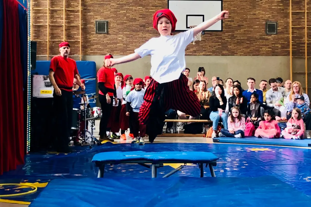 Kind mit rotem Kopftuch und weißem T‑Shirt springt über Sprungtisch in Turnhalle vor Publikum