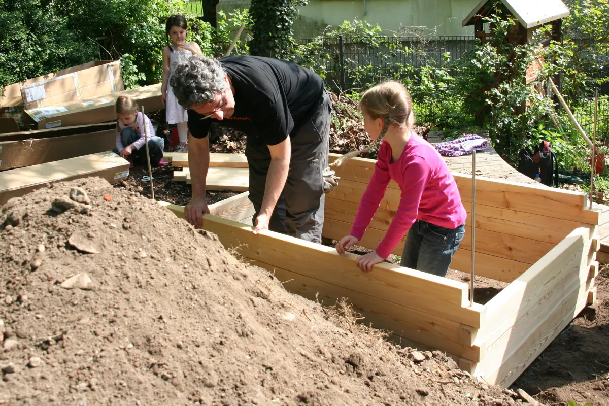 Erwachsener und zwei Kinder bauen ein Holz-Hochbeet im Garten, im Vordergrund großer Erdhügel