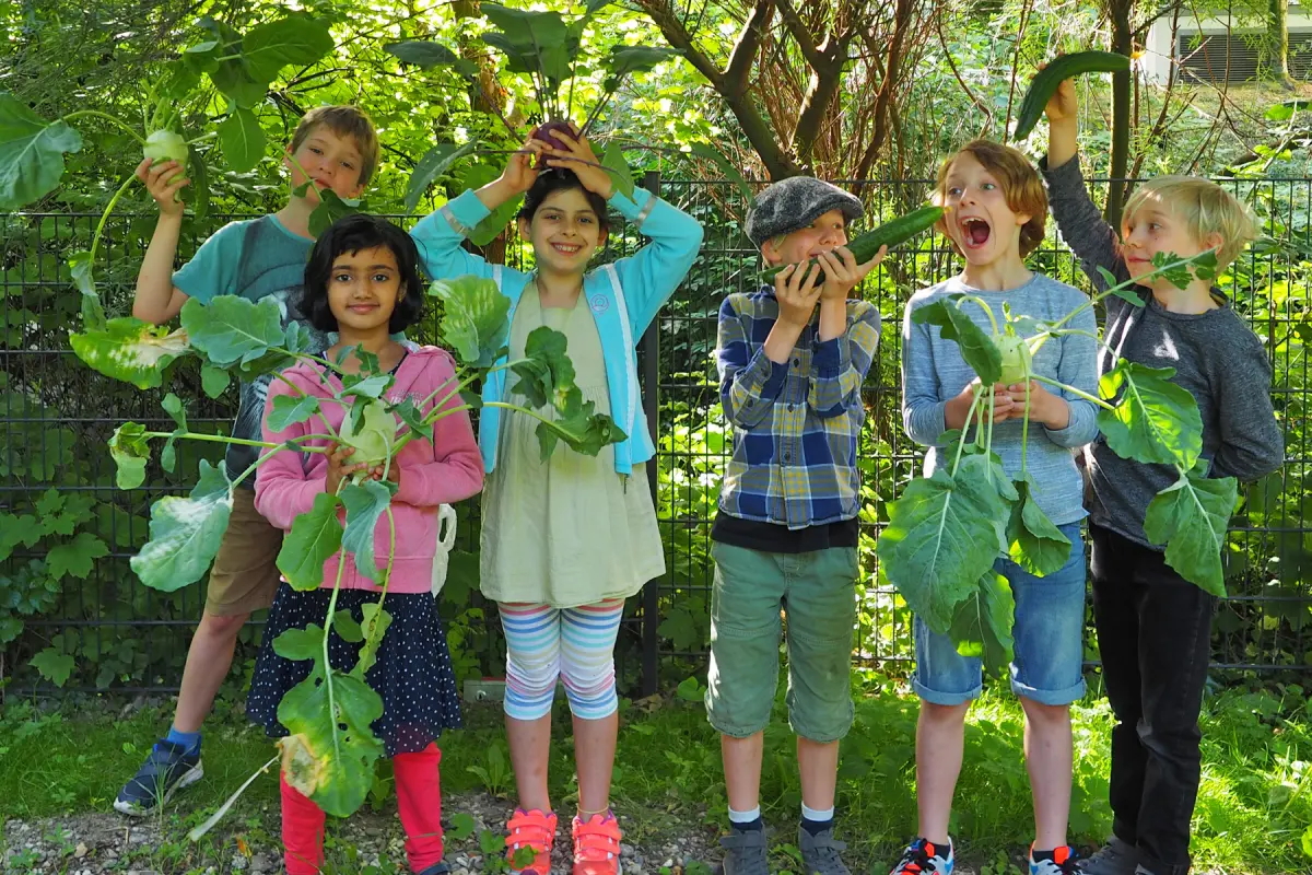 Sechs Kinder stehen vor einem Zaun im Garten und halten große Kohlrabi und Gurken, einige spielen damit.