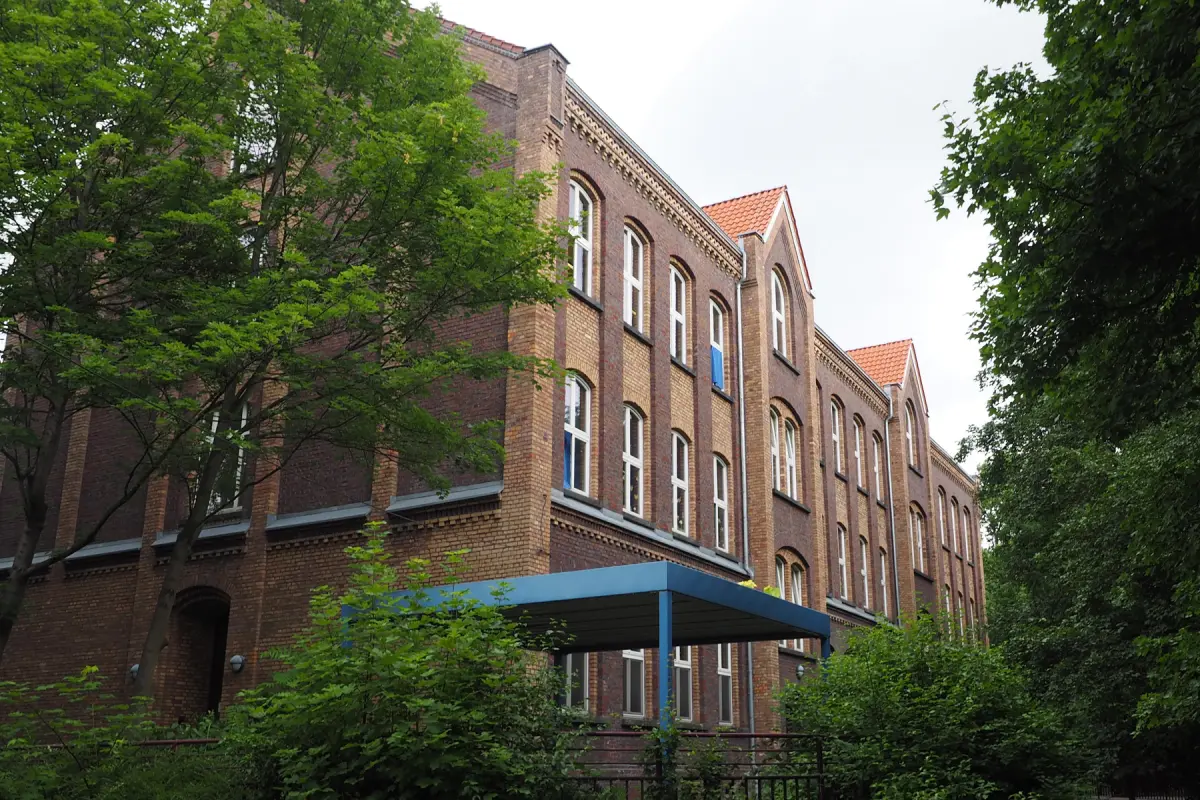Dreigeschossiges rotes Backsteingebäude mit vielen bogenförmigen Fenstern, blauem Vordach und umgebenden Bäumen
