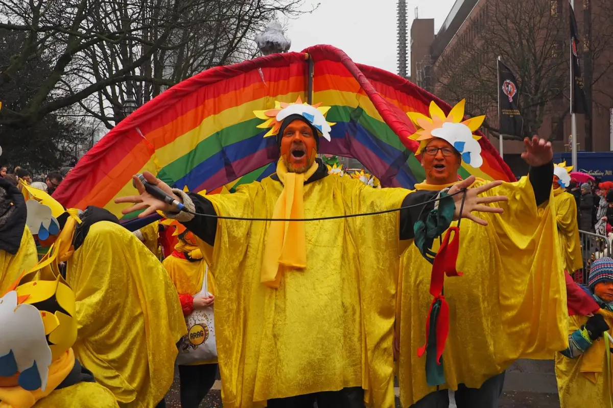 Gruppe verkleideter Menschen in gelben Sonnenkostümen vor großem Regenbogenbanner bei Straßenumzug