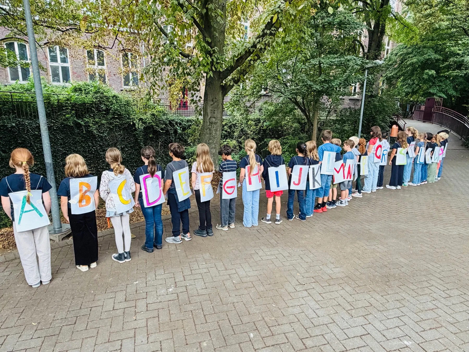 Schulklasse steht in einer Reihe im Freien, Kinder tragen bunte Buchstabenplakate auf dem Rücken