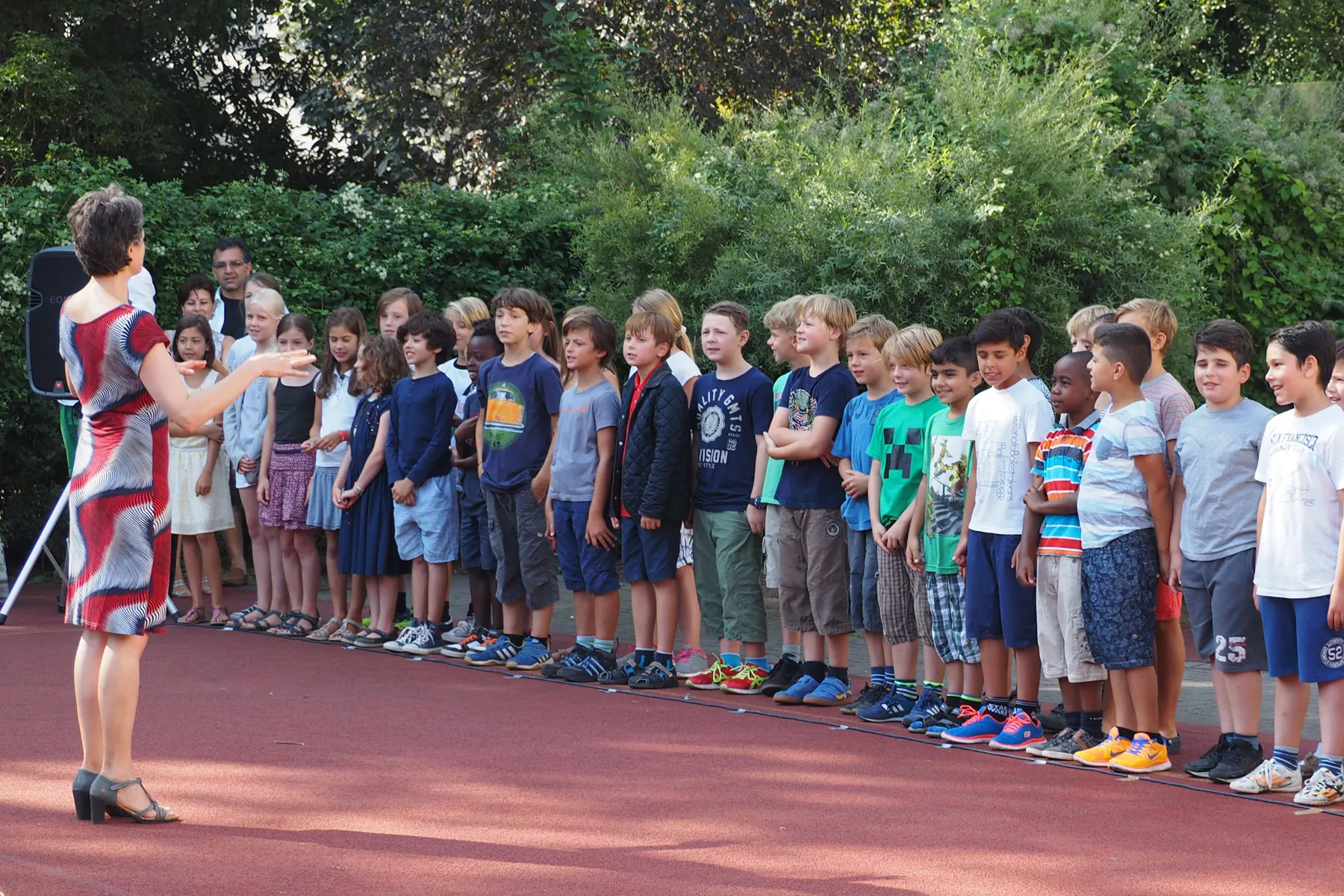Lehrerin dirigiert Reihen von Schulkindern im Freien auf rotem Sportboden vor dichtem Grün