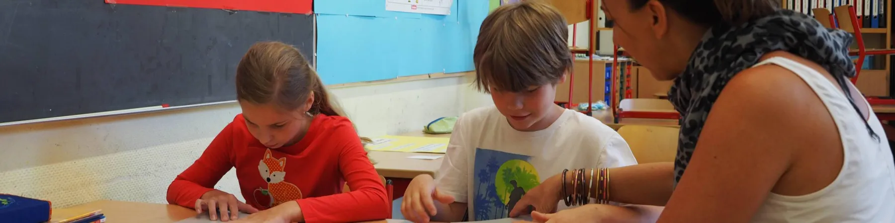Zwei Kinder an Schultisch, Lehrerin zeigt auf Papier; Mädchen in rotem Shirt mit Fuchsmotiv, Junge in weißem T‑Shirt